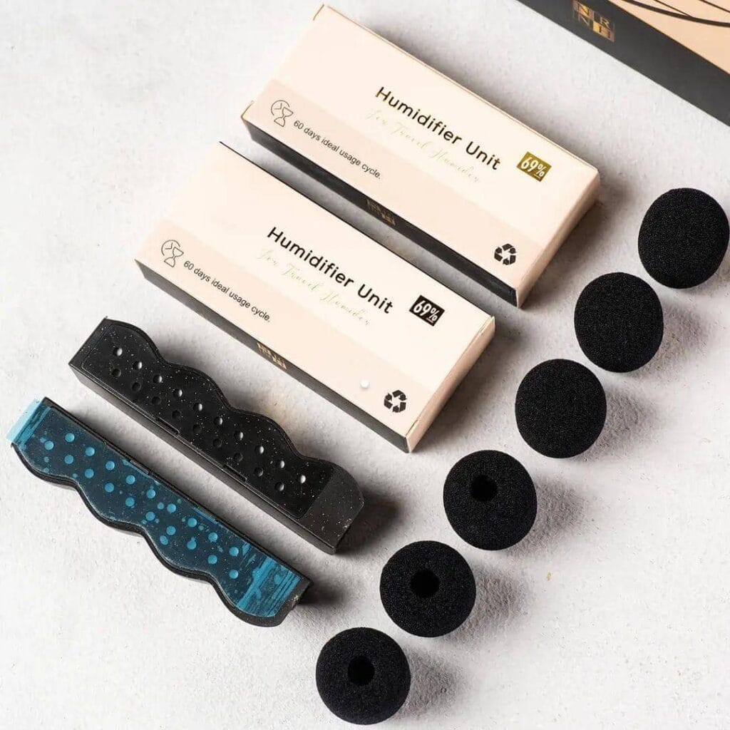 Two beige Galaxy Travel Humidor unit boxes, five black round foam discs, and two rectangular components—one black and one blue with dots, each demonstrating precision engineering—are arranged on a light surface.