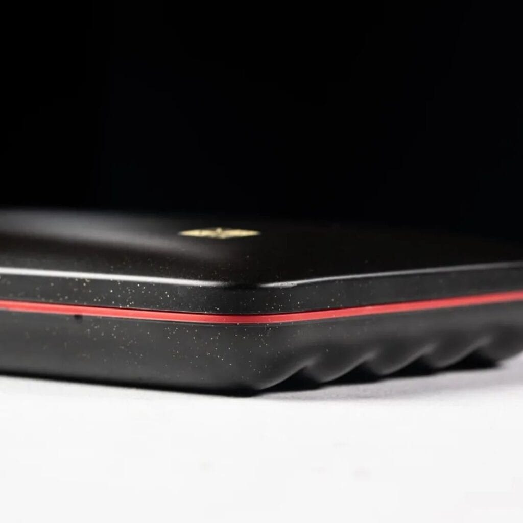 Close-up of a black rectangular object with a red stripe near the edge, textured surface, and a gold logo on top, set against a dark background—expertly crafted with precision engineering inspired by the renowned Galaxy Travel Humidor from the Dominican Republic.