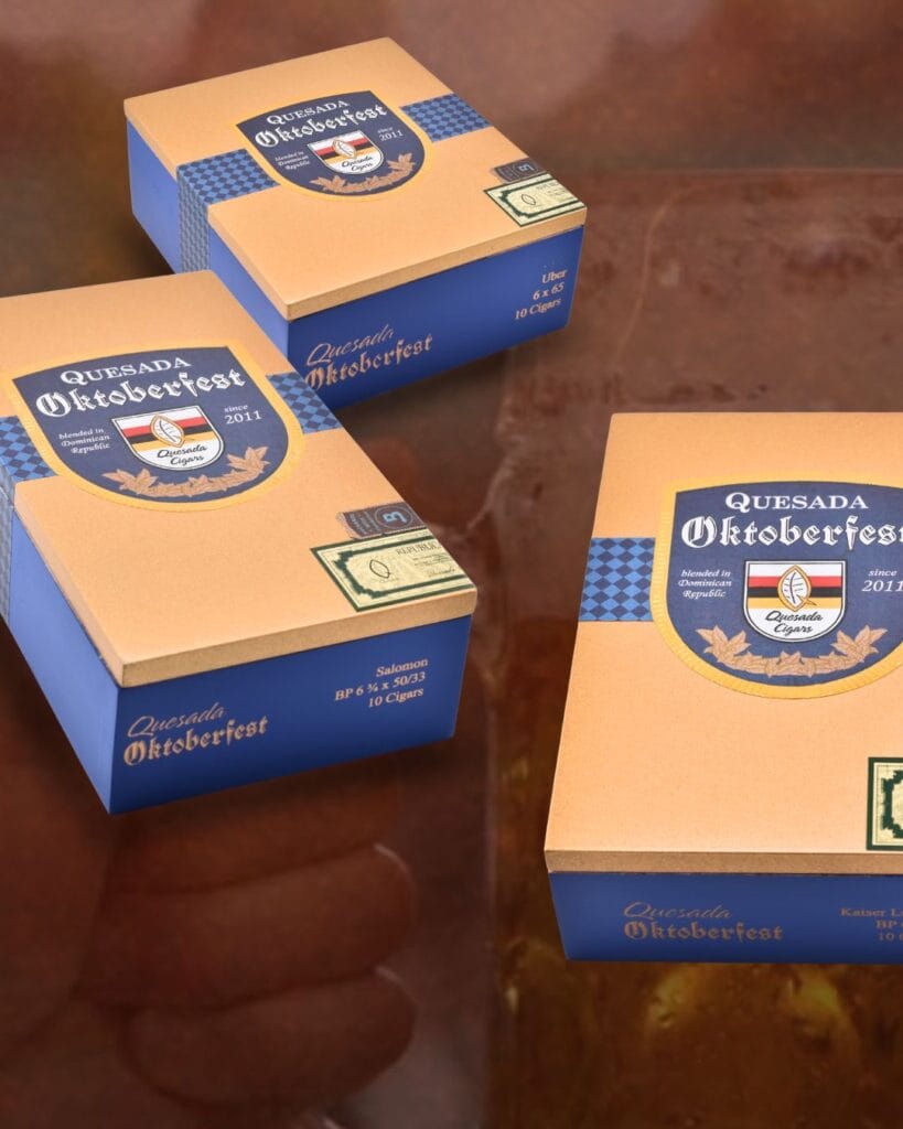 Three orange and blue boxes labeled Quesada Oktoberfest 2026 are displayed on a brown surface. The boxes feature a crest logo and text indicating they contain 10 cigars each, along with a distinctive blue checkered pattern.
