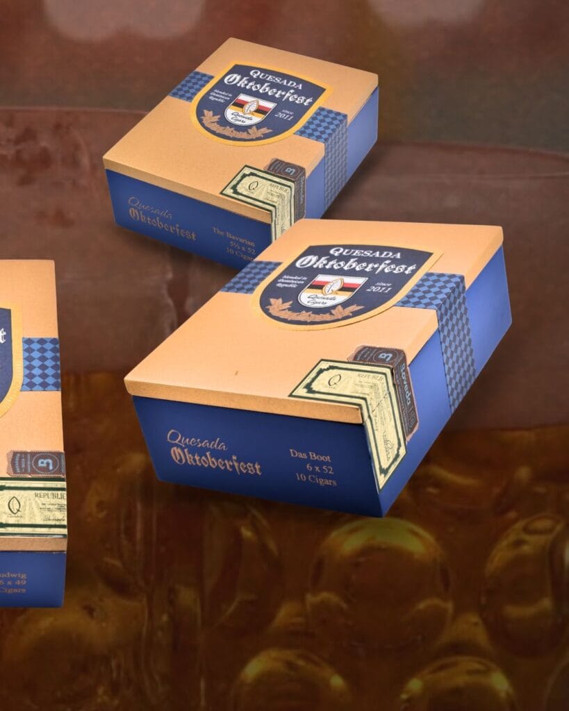 Three orange and blue cigar boxes labeled Quesada Oktoberfest 2026 sit on a blurred brown background, featuring beer stein graphics and decorative accents.